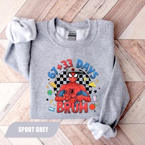 May include: Sport grey sweatshirt featuring a graphic of Spider-Man with the text "67+33 DAYS BRUH". The design includes a checkered background and various cartoon elements. The sweatshirt is made of a soft material.
