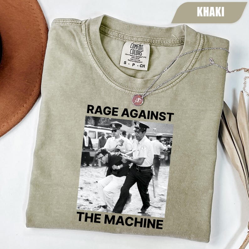 May include: Khaki-colored t-shirt with the words "RAGE AGAINST THE MACHINE" printed in black. The graphic is a black and white image of police officers arresting a person. A silver necklace with a pink pendant rests on the shirt.