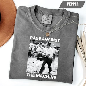 May include: Pepper-colored t-shirt with a black and white image and the text "RAGE AGAINST THE MACHINE". The image depicts a scene of police officers arresting a person. A gold necklace is draped across the shirt.