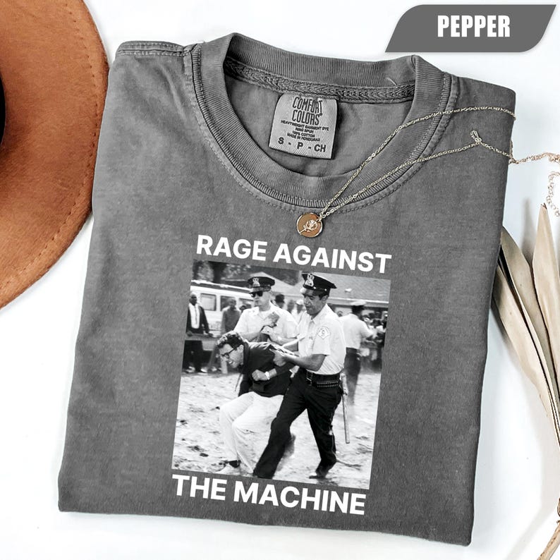 May include: Pepper-colored t-shirt with a black and white image and the text "RAGE AGAINST THE MACHINE". The image depicts a scene of police officers arresting a person. A gold necklace is draped across the shirt.