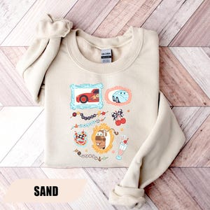 Puede incluir: Una sudadera de cuello redondo color arena con un estampado gr&aacute;fico de personajes de la pel&iacute;cula Cars. El dise&ntilde;o incluye ilustraciones enmarcadas de Rayo McQueen, Sally y Mate, junto con elementos decorativos como cerezas, un batido y texto.