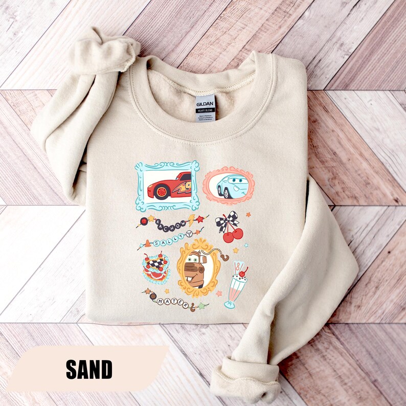 Puede incluir: Una sudadera de cuello redondo color arena con un estampado gr&aacute;fico de personajes de la pel&iacute;cula Cars. El dise&ntilde;o incluye ilustraciones enmarcadas de Rayo McQueen, Sally y Mate, junto con elementos decorativos como cerezas, un batido y texto.