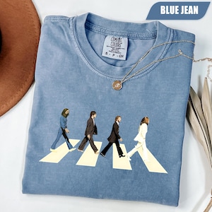 May include: Blue jean coloured t-shirt featuring an Abbey Road graphic. The graphic depicts four figures walking across a crosswalk. The shirt has a crew neck and short sleeves. A silver necklace with a pendant is draped across the shirt.