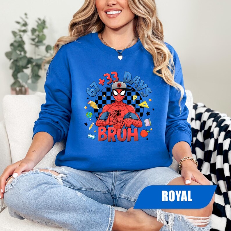 May include: Royal blue sweatshirt featuring a graphic of Spider-Man with the text "67 + 33 DAYS BRUH". The design includes a checkered background, stars, and school-themed elements. The sweatshirt is a long-sleeved crew neck.