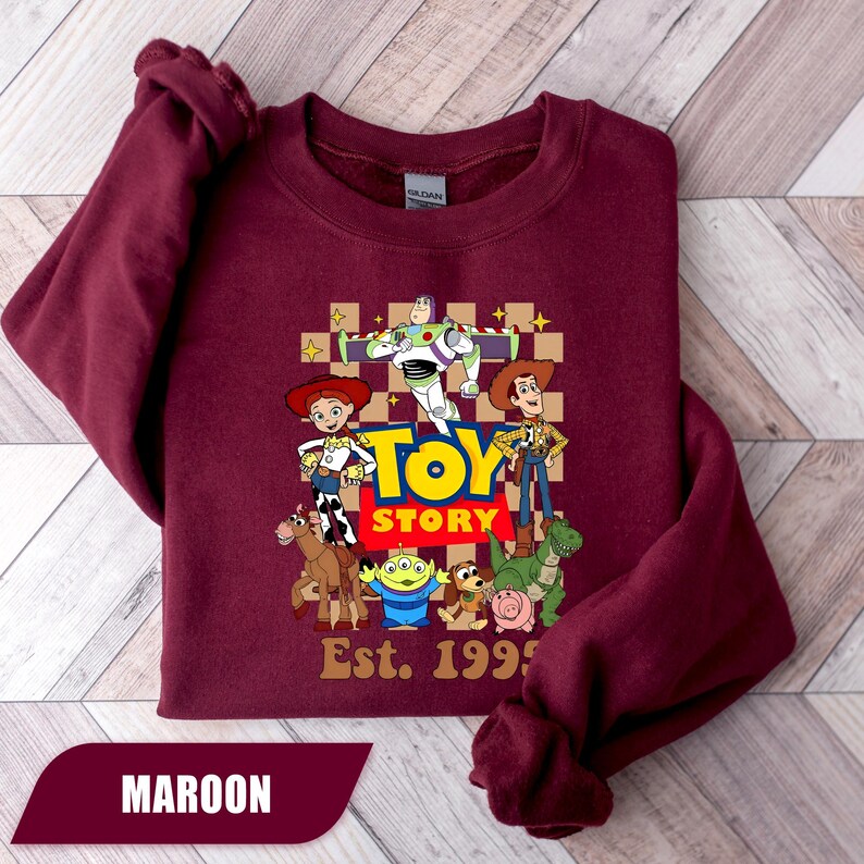 Puede incluir: Sudadera granate con un gr&aacute;fico de los personajes de Toy Story, incluyendo a Woody, Buzz Lightyear y Jessie. Las palabras "Toy Story" est&aacute;n en amarillo y rojo, con "Est. 1995" debajo. La palabra "MAROON" est&aacute; en la parte inferior.