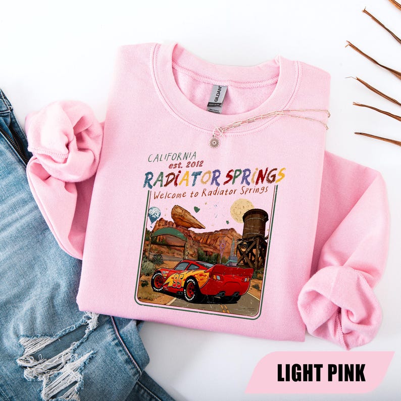 May include: A light pink crewneck sweatshirt featuring a graphic print of a red race car with the number 95. The text reads California est. 2012, Radiator Springs, and Welcome to Radiator Springs, set against a desert landscape with a water tower.