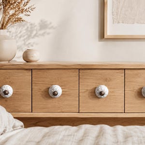 May include: Four round white ceramic cabinet knobs with intricate embossed floral patterns and dark metallic centres, each mounted on a light oak wood drawer front. The knobs feature a textured surface with a repeating petal design.