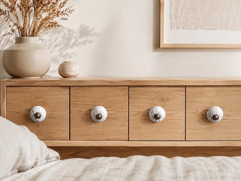May include: Four round white ceramic cabinet knobs with intricate embossed floral patterns and dark metallic centres, each mounted on a light oak wood drawer front. The knobs feature a textured surface with a repeating petal design.