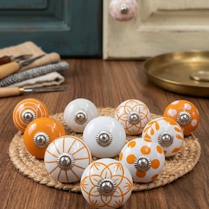 Pode incluir: Uma coleção de puxadores de armário de cerâmica decorativos em vários designs e cores, incluindo branco e laranja. Os puxadores apresentam padrões florais e geométricos com ferragens de metal prateado. Dispostos sobre uma esteira tecida.