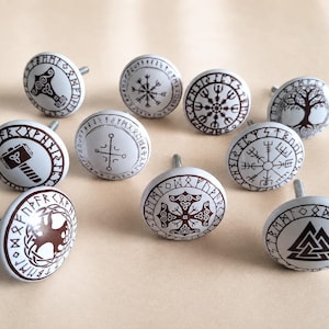 May include: A set of ten round white ceramic cabinet knobs, each featuring a unique dark brown printed design including Norse symbols, runes, a tree of life, a hammer, and geometric patterns, with a metal screw extending from the back of each.