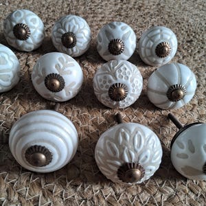 May include: A collection of ten round ceramic knobs featuring various embossed patterns in off-white and beige tones. Each knob is centred with a small, circular, antique-style bronze metal hardware piece and includes a threaded metal bolt on the back.