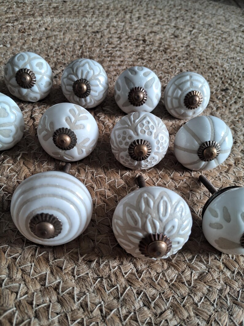 May include: A collection of ten round ceramic knobs featuring various embossed patterns in off-white and beige tones. Each knob is centred with a small, circular, antique-style bronze metal hardware piece and includes a threaded metal bolt on the back.