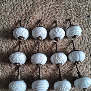 May include: A collection of twelve round cabinet knobs featuring white ceramic surfaces with embossed beige patterns. Each knob is equipped with a threaded metal bolt and a bronze-toned base, showcasing various geometric and floral designs.