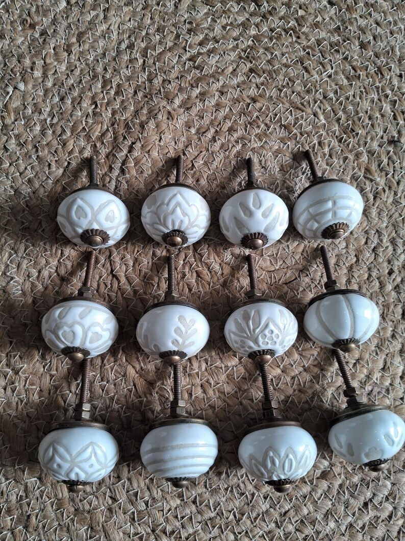 May include: A collection of twelve round cabinet knobs featuring white ceramic surfaces with embossed beige patterns. Each knob is equipped with a threaded metal bolt and a bronze-toned base, showcasing various geometric and floral designs.