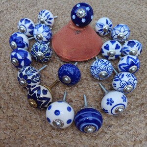 May include: A selection of ceramic cabinet knobs with blue and white patterns. The knobs display floral, polka dot, and striped designs. Each knob has a metal screw. The knobs are arranged on a woven surface, creating a decorative display.