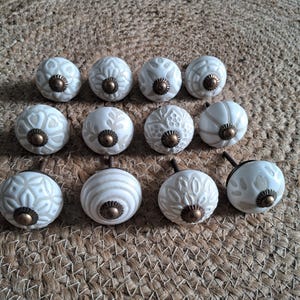 May include: A collection of twelve round ceramic cabinet knobs featuring various off-white and beige embossed patterns. Each knob is finished with a small, circular, antique-style brass-toned metal centre piece and a matching metal screw base.