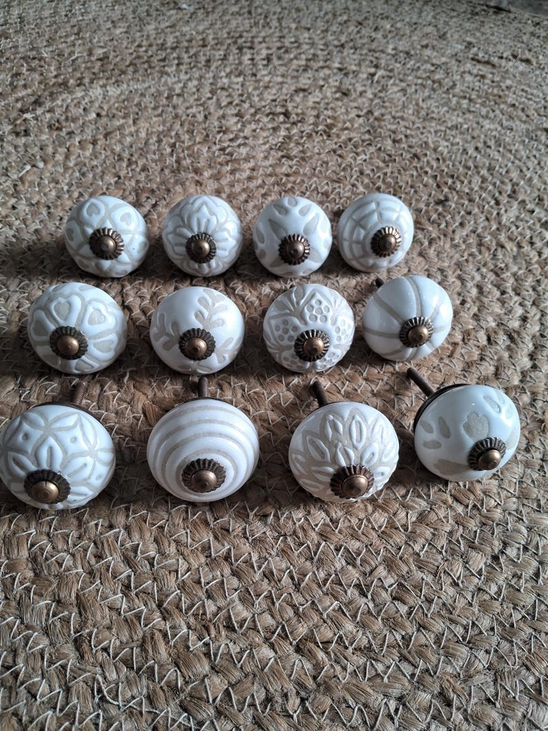 May include: A collection of twelve round ceramic cabinet knobs featuring various off-white and beige embossed patterns. Each knob is finished with a small, circular, antique-style brass-toned metal centre piece and a matching metal screw base.