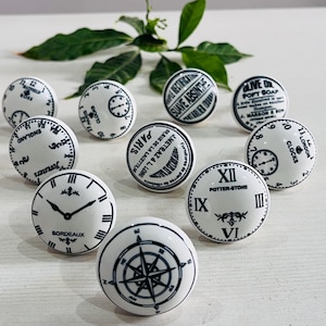 May include: A selection of white ceramic cabinet knobs with black designs. The knobs display clock faces, a compass rose, and text including "BORDEAUX", "PARIS", and "POTTER-STONE". Green leaves are in the background.