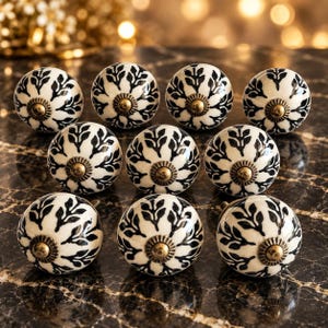 May include: A set of ten round cabinet knobs featuring a white base with a black leaf pattern radiating from the centre. Each knob has a central antique brass-toned metal hardware piece with a ridged circular design at the point of attachment.