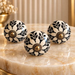 May include: Three round ceramic knobs featuring a cream-colored base with a black hand-painted leaf pattern. Each knob has a central antique brass-toned metal hardware piece with a fluted edge, providing a decorative accent to the spherical design.