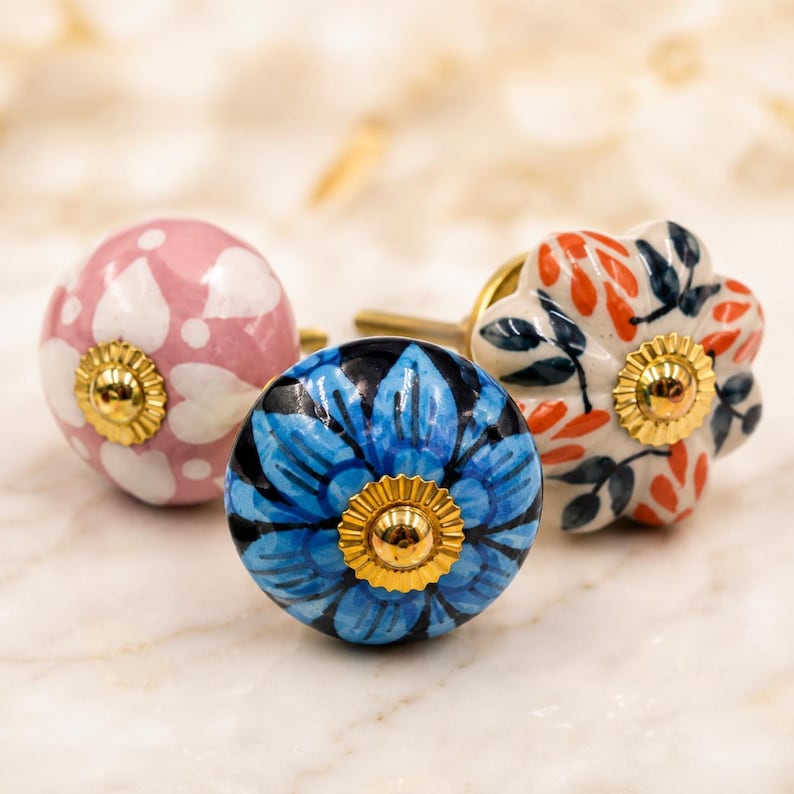 Pode incluir: Tr&ecirc;s puxadores redondos de cer&acirc;mica para arm&aacute;rios com padr&otilde;es florais. Um puxador &eacute; rosa com p&eacute;talas brancas, um &eacute; azul com p&eacute;talas azul-escuras e um &eacute; branco com folhas laranja e azul-escuras. Cada puxador tem uma pe&ccedil;a central de metal dourado.