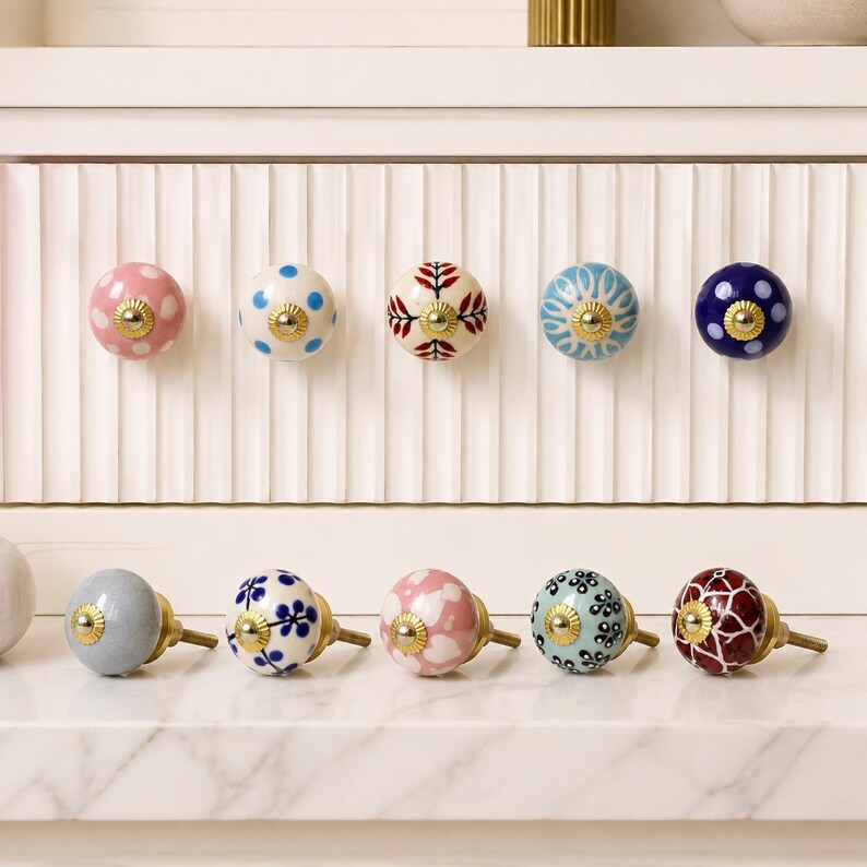 Pode incluir: Uma cole&ccedil;&atilde;o de dez puxadores redondos de cer&acirc;mica para arm&aacute;rios com v&aacute;rios padr&otilde;es, incluindo bolinhas, motivos florais e designs geom&eacute;tricos em cores como rosa, azul, branco e vermelho escuro, com uma pe&ccedil;a central de metal dourado.