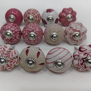 Op de afbeelding: Een verzameling van twaalf ronde keramische kastknoppen met diverse roze, witte en beige patronen. Elke knop heeft een centrale zilverkleurige metalen schroef en een decoratieve metalen ring aan de voorzijde, met bloem-, streep- en geometrische ontwerpen.