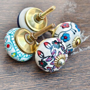 Pu&ograve; includere: Quattro pomelli decorativi in ceramica per mobili con ferramenta in ottone. I pomelli presentano disegni floreali e geometrici dipinti a mano in vari colori, tra cui blu, rosso e verde, su sfondo bianco. I pomelli sono montati su una superficie di legno.