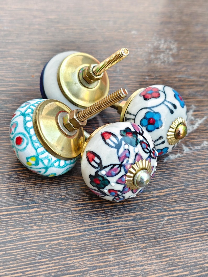 Pu&ograve; includere: Quattro pomelli decorativi in ceramica per mobili con ferramenta in ottone. I pomelli presentano disegni floreali e geometrici dipinti a mano in vari colori, tra cui blu, rosso e verde, su sfondo bianco. I pomelli sono montati su una superficie di legno.