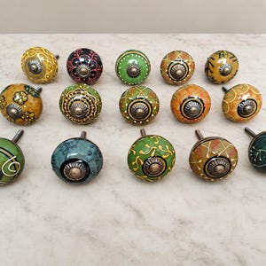 May include: A collection of fourteen round ceramic cabinet knobs featuring various hand-painted patterns in shades of green, yellow, orange, and black. Each knob has a central metallic hardware piece and a smooth, glossy finish with intricate designs.