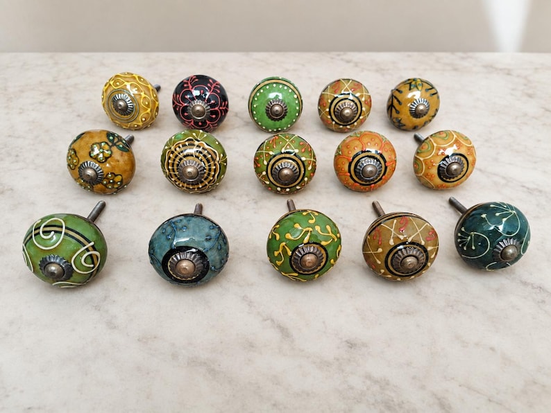 May include: A collection of fourteen round ceramic cabinet knobs featuring various hand-painted patterns in shades of green, yellow, orange, and black. Each knob has a central metallic hardware piece and a smooth, glossy finish with intricate designs.