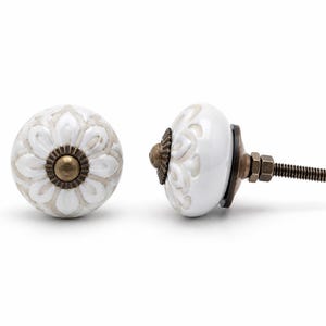 May include: Two round cabinet knobs featuring a white ceramic surface with a raised, light beige floral pattern. Each knob has a central antique brass-toned metal cap and a matching metal base with a threaded bolt and nut for installation.