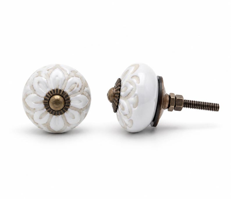 May include: Two round cabinet knobs featuring a white ceramic surface with a raised, light beige floral pattern. Each knob has a central antique brass-toned metal cap and a matching metal base with a threaded bolt and nut for installation.
