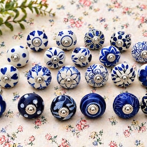 May include: A collection of decorative ceramic cabinet knobs in various designs. The knobs feature white and blue floral, heart, and geometric patterns. Each knob has a silver-colored metal base and stem. The knobs are arranged on a floral patterned surface.