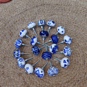 Peut inclure: Ensemble de boutons de meubles en céramique bleus et blancs, avec des motifs floraux et géométriques variés. Les boutons sont disposés en cercle. Chaque bouton est équipé d'une vis métallique. Fond texturé de couleur neutre.