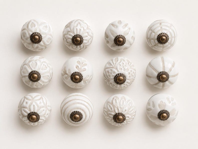 May include: A set of twelve round ceramic cabinet knobs arranged in a grid. Each knob features a white base with light grey embossed patterns and a central antique brass-toned metal hardware piece with a scalloped edge and a small rounded centre.