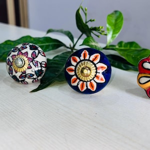 Può includere: Quattro pomelli decorativi in ceramica con disegni floreali. I pomelli sono bianchi, blu e blu navy, con accenti rossi, arancioni e gialli. Ogni pomello ha una base in metallo color oro e un motivo floreale unico.