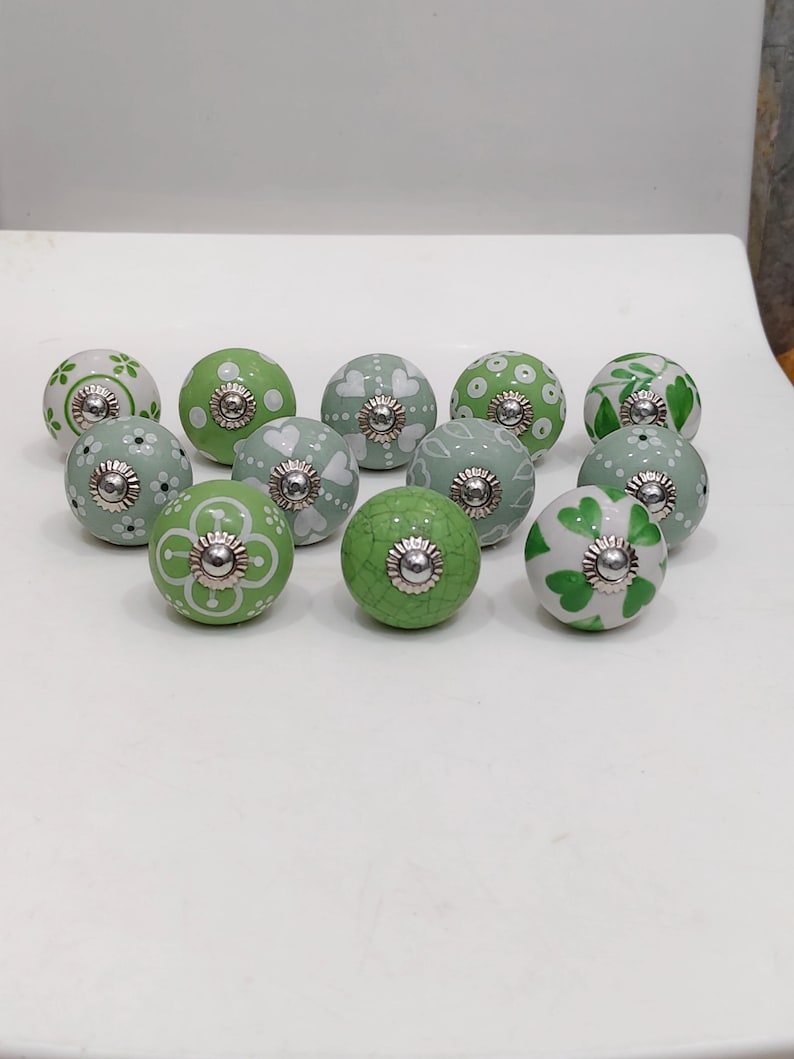 Pode incluir: Uma cole&ccedil;&atilde;o de puxadores de arm&aacute;rio de cer&acirc;mica decorativos em v&aacute;rios tons de verde e branco. Cada puxador apresenta um design pintado &uacute;nico, incluindo padr&otilde;es florais, cora&ccedil;&otilde;es e pontos. Os puxadores t&ecirc;m uma base de metal prateado.