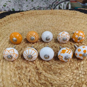 May include: A set of ten round ceramic cabinet knobs featuring various orange and white patterns, including solid orange, white polka dots, orange floral motifs, and orange striped designs, each with a central silver-toned metal screw hardware.