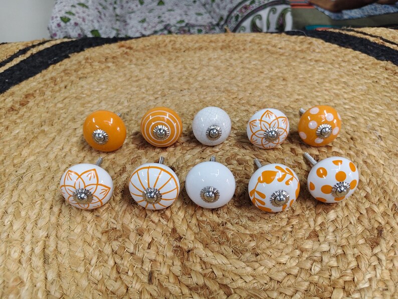 May include: A set of ten round ceramic cabinet knobs featuring various orange and white patterns, including solid orange, white polka dots, orange floral motifs, and orange striped designs, each with a central silver-toned metal screw hardware.