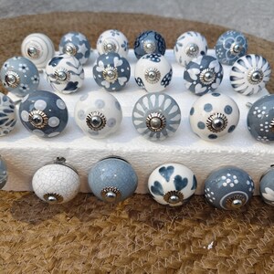 May include: A selection of ceramic cabinet knobs in blue and white. The knobs display diverse patterns, including floral and polka dots, with silver-coloured metal fixings. They are arranged on a white surface, offering a decorative touch.