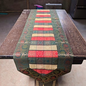 May include: A decorative table runner with a detailed floral pattern in shades of green, red, and gold. The runner features a central panel with alternating stripes of red, gold, and green. The runner is approximately 152 cm long.