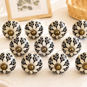 May include: A set of eleven round cabinet knobs featuring a white base with a black leaf-like pattern radiating from the center. Each knob is finished with a small, circular, antique brass-toned metal hardware piece at the very center point.