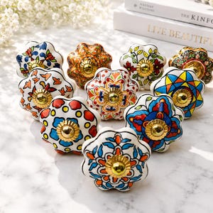 Op de afbeelding: Een verzameling van tien keramische ladeknoppen met geschulpte randen en een glanzende afwerking. Elke knop heeft een uniek handgeschilderd bloemen- of geometrisch patroon in levendige kleuren zoals blauw, oranje, rood en groen, met een goudkleurig metalen beslag in het midden.