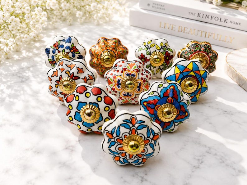 Op de afbeelding: Een verzameling van tien keramische ladeknoppen met geschulpte randen en een glanzende afwerking. Elke knop heeft een uniek handgeschilderd bloemen- of geometrisch patroon in levendige kleuren zoals blauw, oranje, rood en groen, met een goudkleurig metalen beslag in het midden.