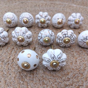 May include: A collection of eleven white ceramic cabinet knobs with gold-colored accents. The knobs feature various floral and geometric designs, with gold-colored hardware. The knobs are arranged on a woven, beige surface.