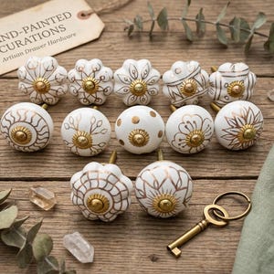 Peut inclure: Ensemble de boutons de tiroir en céramique peints à la main, blancs et dorés, avec divers motifs floraux et géométriques. Une clé en laiton et une étiquette "Hand-Painted Curations Artisan Drawer Hardware" sont également présentes. Les boutons sont disposés sur une surface en bois.