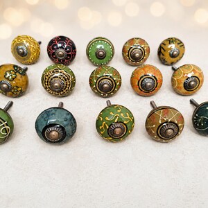 May include: A collection of fourteen round ceramic cabinet knobs arranged in three rows. Each knob features a unique hand-painted pattern in shades of green, yellow, orange, and black, finished with a central metallic bolt and a glossy surface.