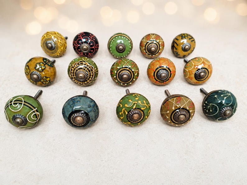 May include: A collection of fourteen round ceramic cabinet knobs arranged in three rows. Each knob features a unique hand-painted pattern in shades of green, yellow, orange, and black, finished with a central metallic bolt and a glossy surface.