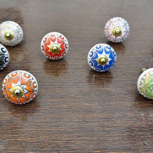 Pu&ograve; includere: Una collezione di sette pomelli rotondi in ceramica per mobili con intricati motivi floreali multicolori nelle tonalit&agrave; arancione, rosso, blu, verde, nero e bianco, ciascuno centrato con un piccolo dettaglio in metallo color oro.